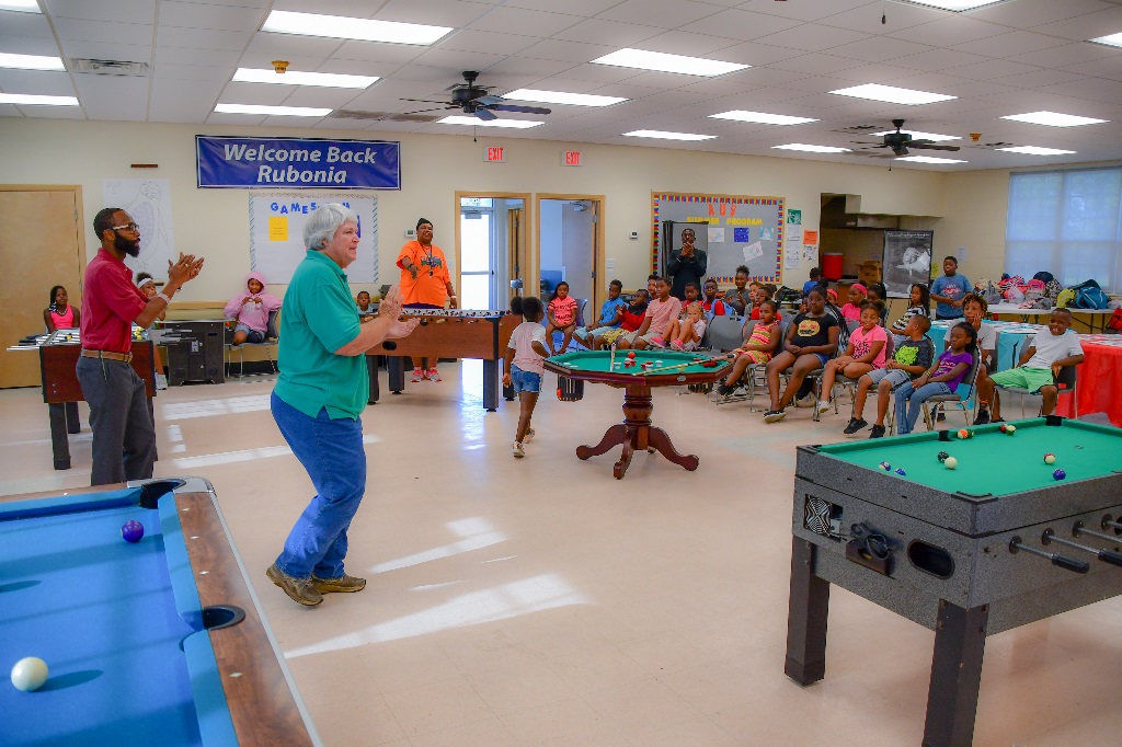 Youth programming at the Rubonia Community Center. Image depicts instructors standing and speaking to seated children, in the main meeting room. There are recreation equipment such as pool tables and foosball tables in the room.
