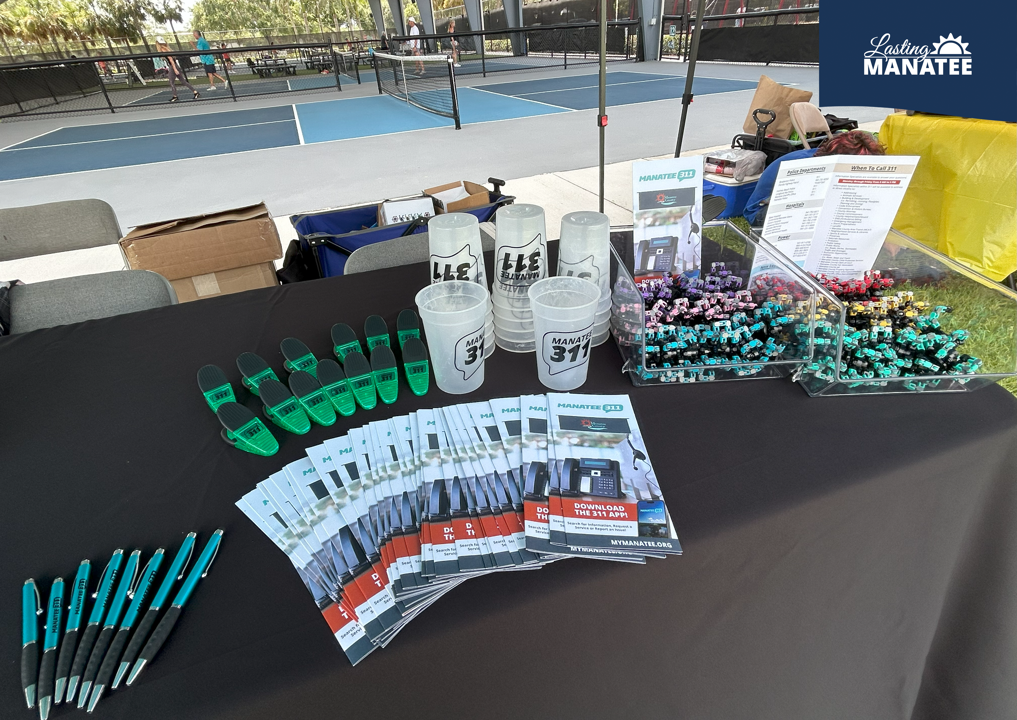 Fidget spinner toys, Manatee 311 pamphlets, and Manatee 311 branded items (pens, cups, chip clips) on top of vendor table at G.T. Bray Park for Family Fun Night - August 1, 2025