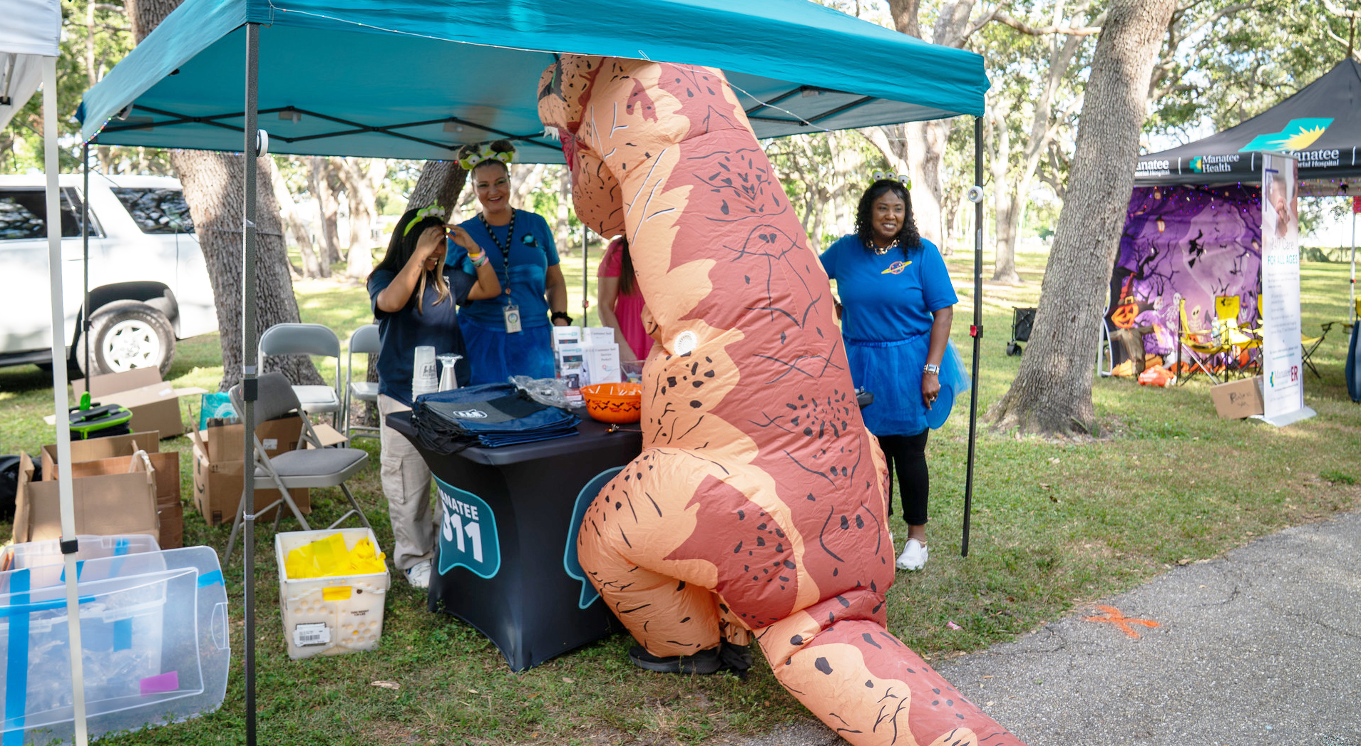Attendee in a large inflatable T-Rex costume leans over the Manatee 311 booth at an outdoor community event, engaging with staff under a blue canopy. There is a nearby tent with decorations. The table has Halloween candy and branded 311 merchandise
