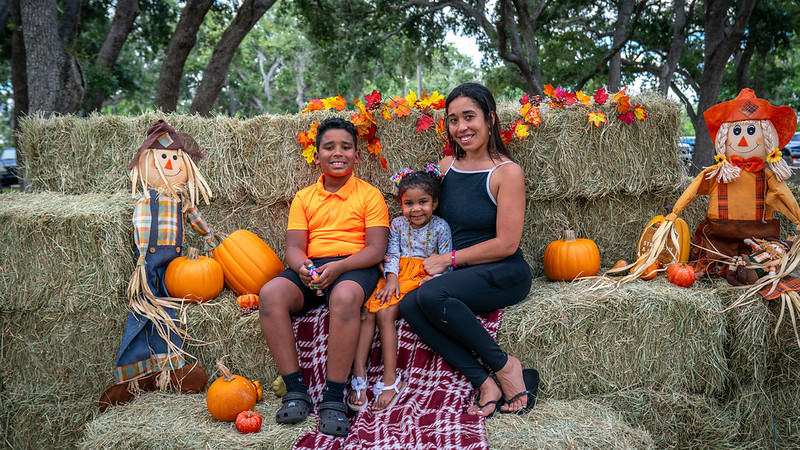 A mother and her two smiling children pose for a photo at an outdoor autumn event, sitting on hay bales decorated with scarecrows, pumpkins, and artificial fall leaves.