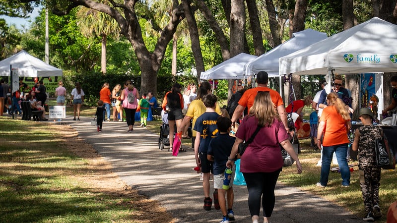 A park pathway lined with vendors under white canopy tents, crowded with diverse people strolling between the booths on a sunny day.