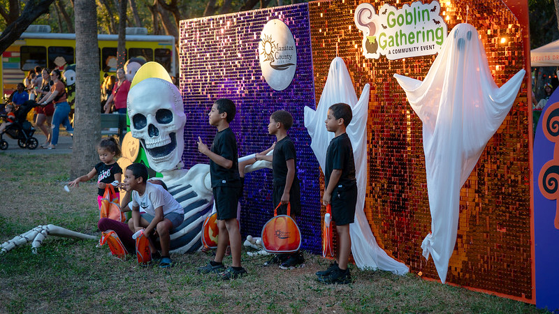 An outdoor Halloween event photo area with a large skeleton decoration and shiny purple and orange sequin backdrop. Four young boys in black shirts are posing for a photo near hanging white ghost decorations and a