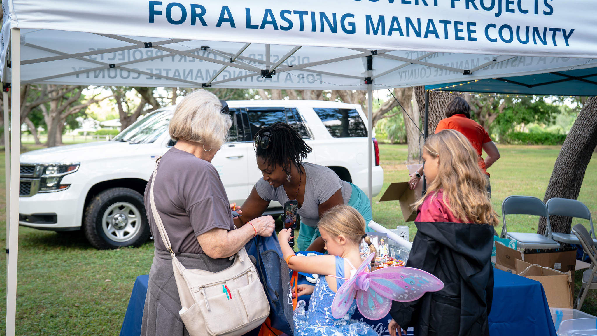 An outdoor event booth features an older woman engaging with a staff member and two children, one in a blue fairy costume with butterfly wings. A white SUV is parked behind the booth, which is under a canopy tent with