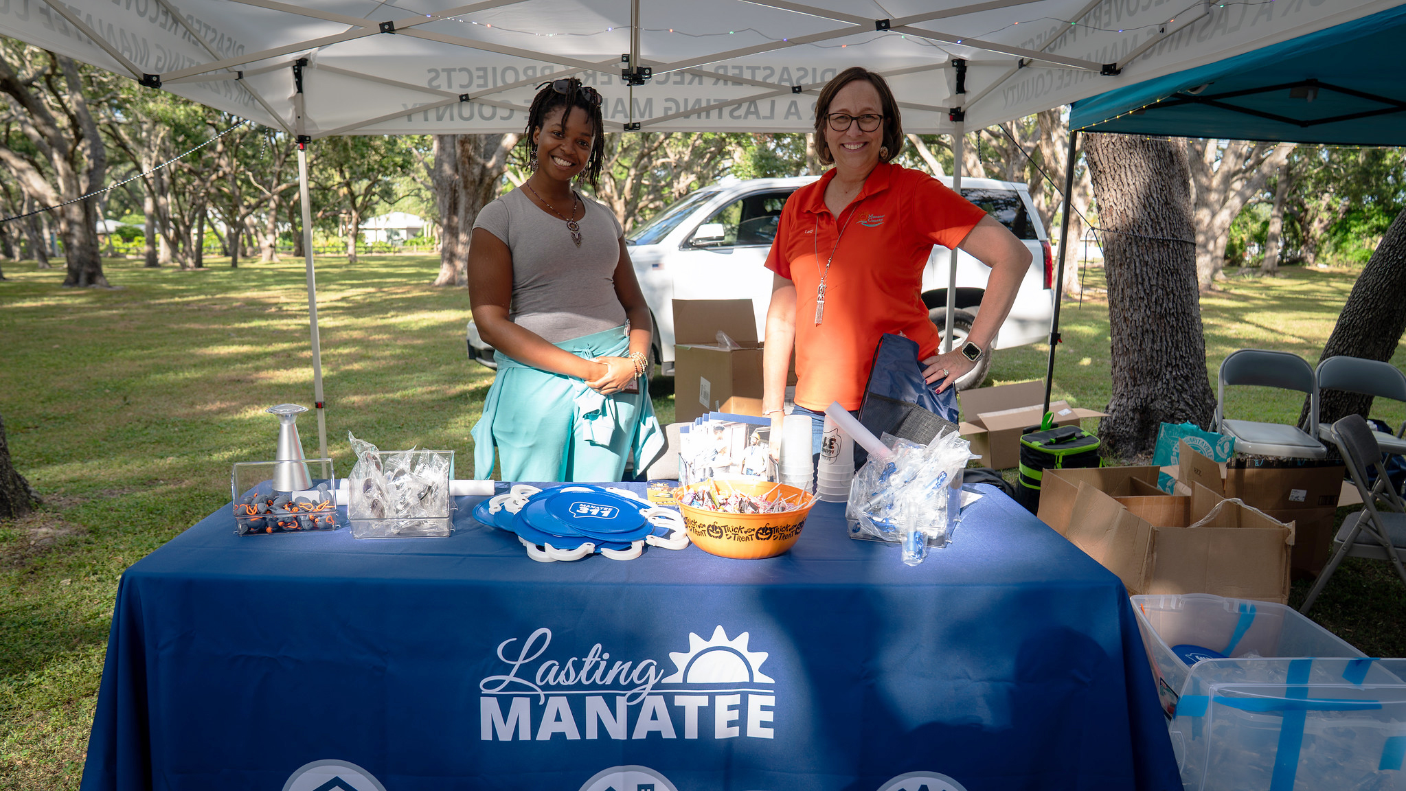 An outdoor Lasting Manatee event booth under a white canopy features two smiling staff members. The table has an orange candy bowl, Lasting Manatee program pamphlets, and different giveaway toys and branded goods.