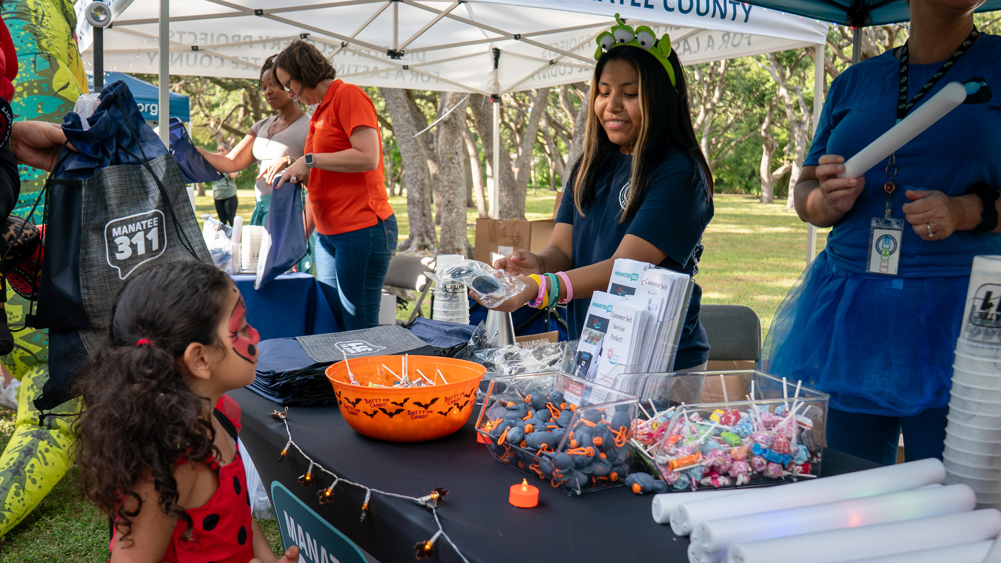 An outdoor event booth features several people, including an adult handing an item to a child in a ladybug costume. The table has an orange jack-o'-lantern candy bowl, stacks of materials, and small gray favors, with a