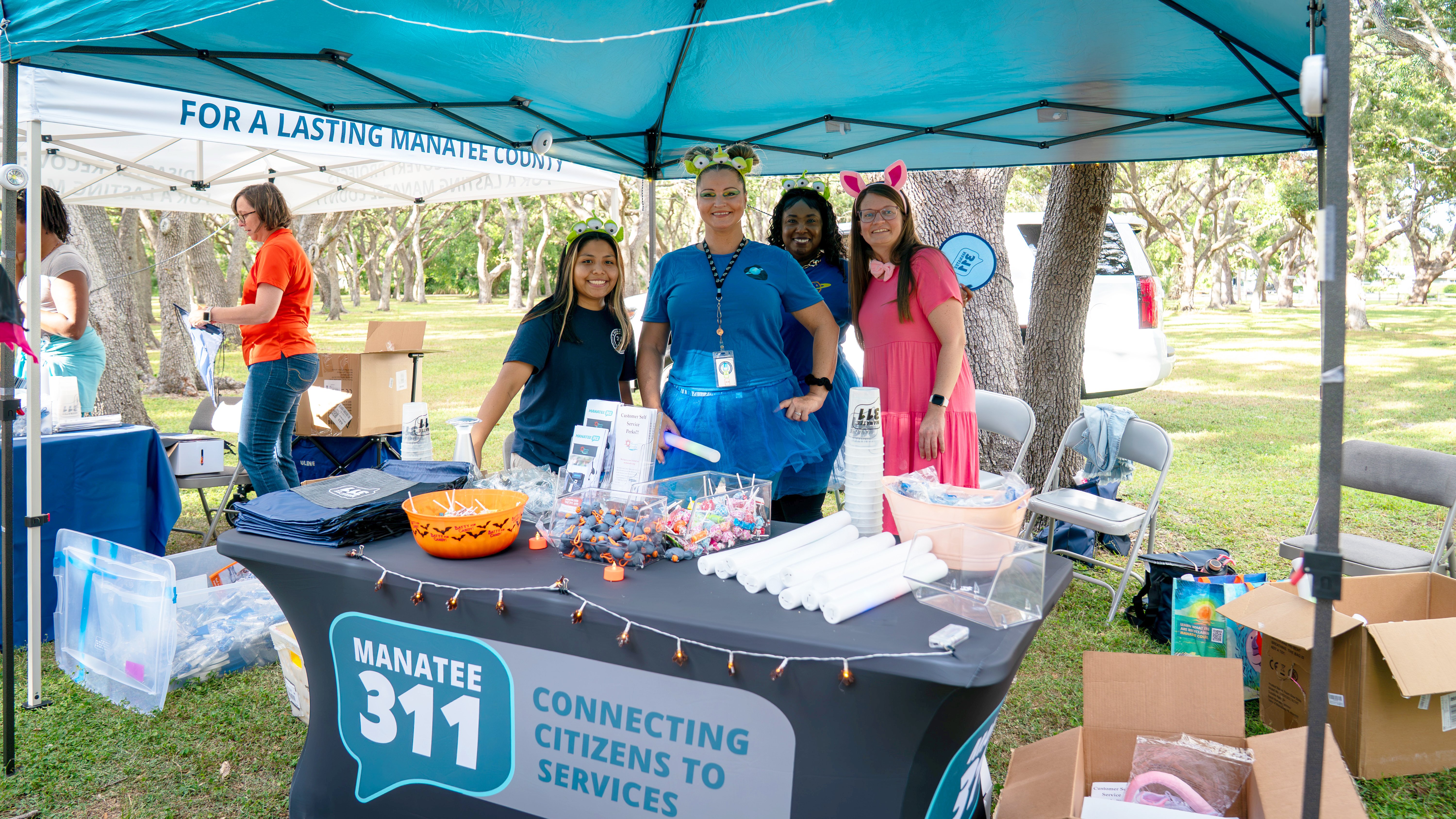 Lasting Manatee and Manatee 311 at Goblin Gathering An outdoor Manatee 311 event booth under a teal canopy features several smiling staff members. The table has an orange candy bowl, various program pamphlets, and different giveaway toys and branded goods.
