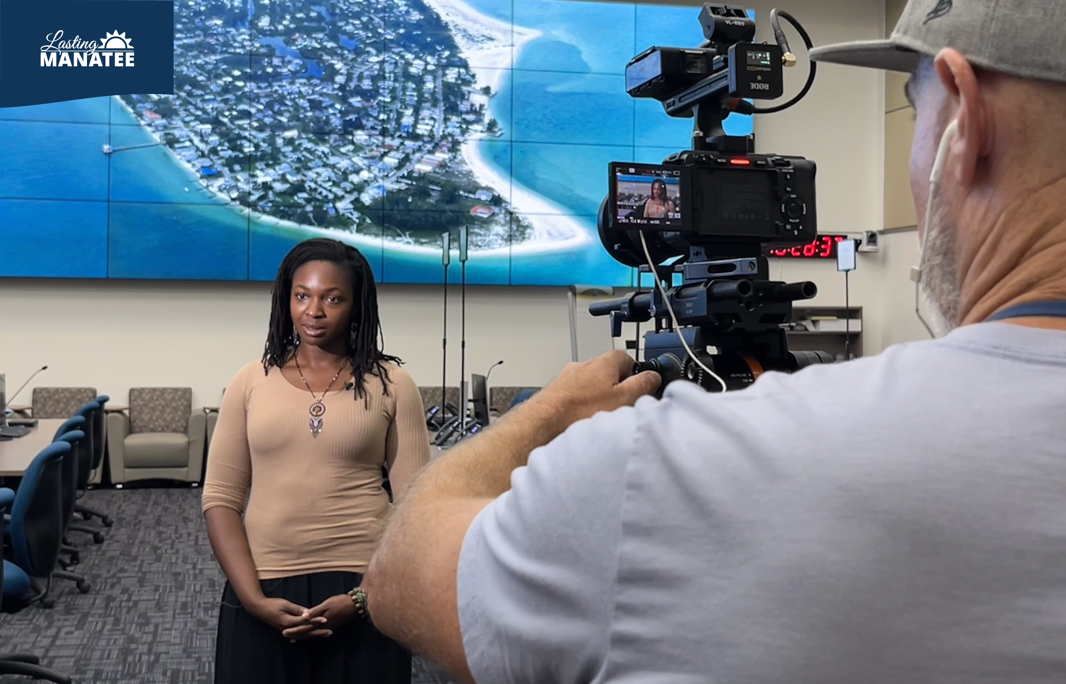 Lasting Manatee interviewed by FOX13 at the Emergency Operations Center Inside the Manatee County Emergency Operations Center, a staff member of Lasting Manatee speaks on camera while FOX13's cameraman films from the right side. The room has desks, chairs, and monitors, with a large TV screen on the back wall.