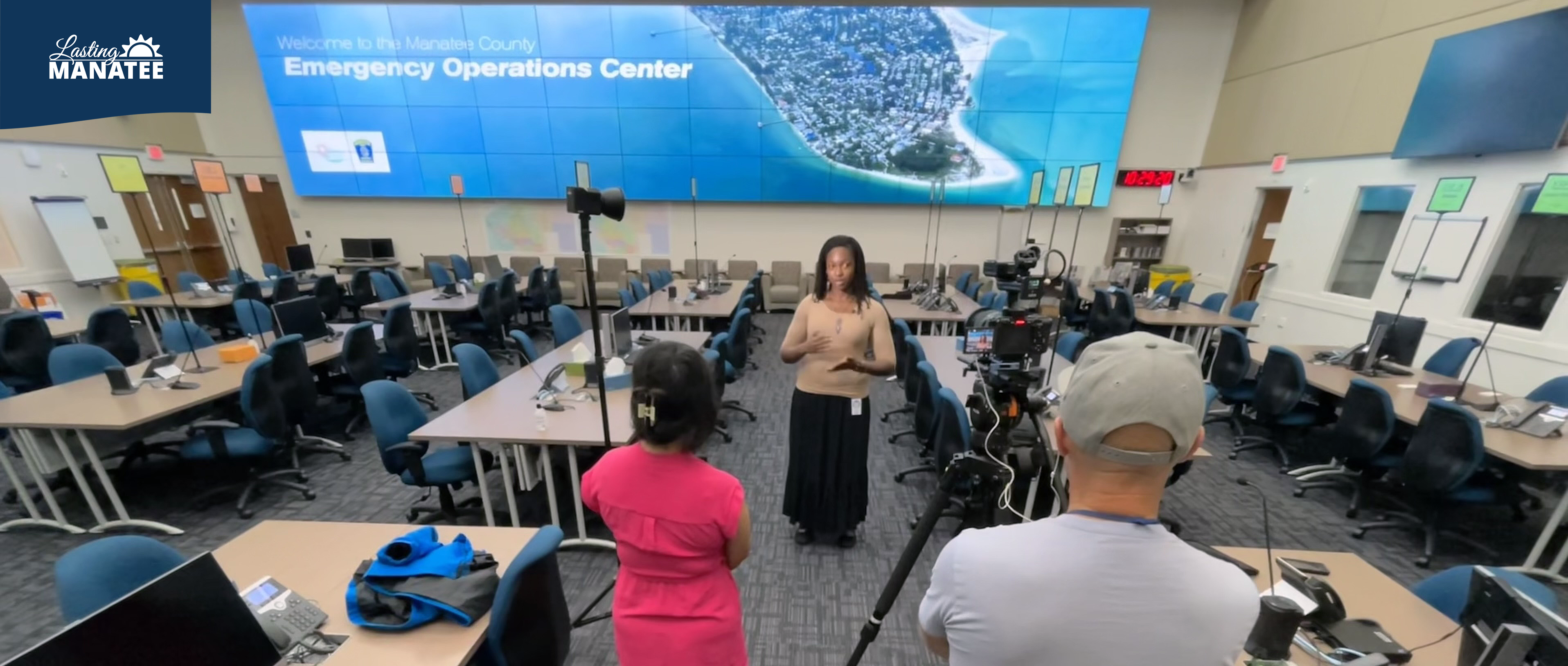 Lasting Manatee interviewed by FOX13 Kimberly Kuizon at the Emergency Operations Center Inside the Manatee County Emergency Operations Center, a staff member of Lasting Manatee speaks on camera while FOX13's reporter holds a microphone while their cameraman films. The room has desks and monitors, with a large TV screen on the back wall.