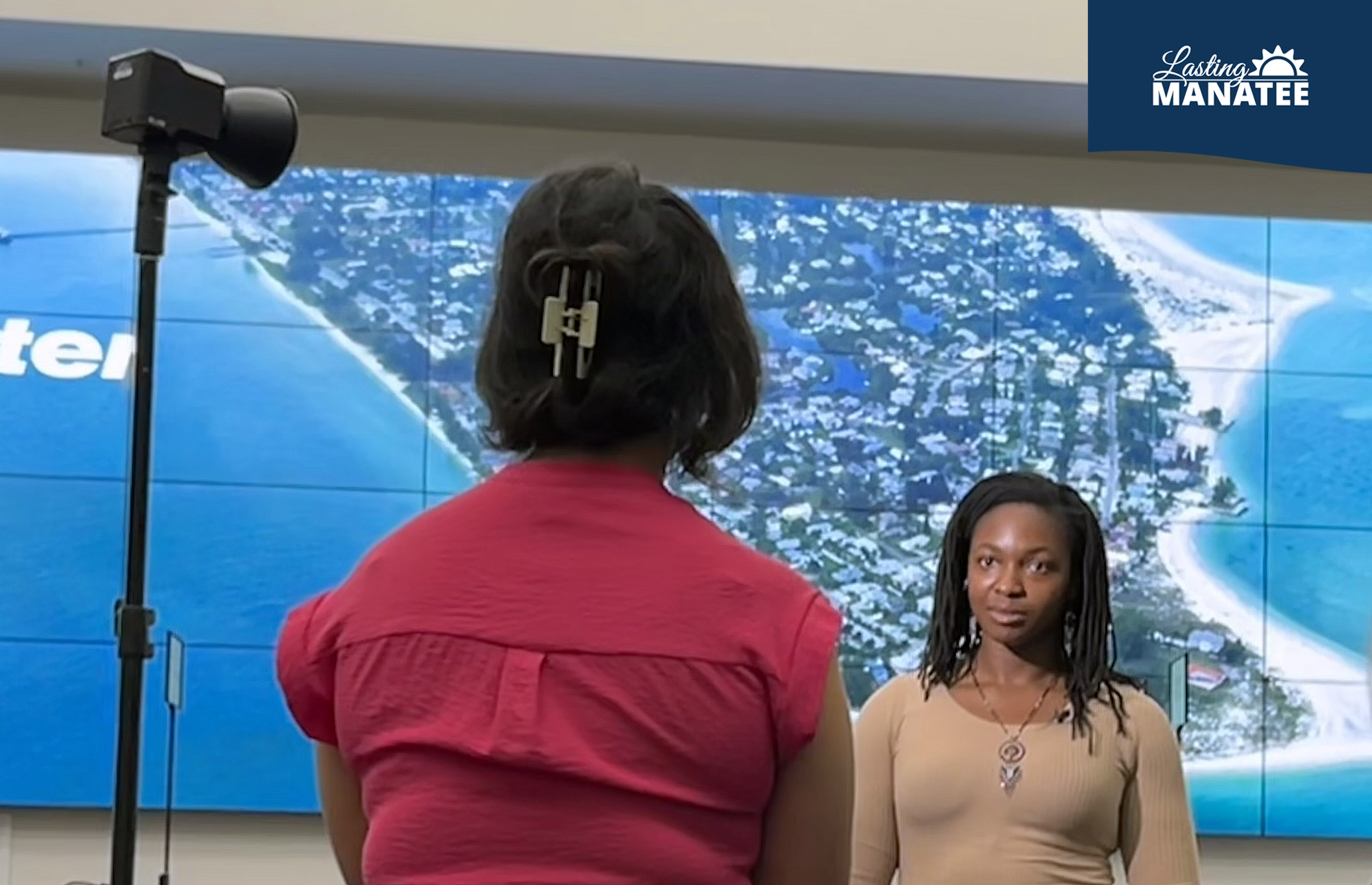 Lasting Manatee interviewed by FOX13 Kimberly Kuizon at the Emergency Operations Center Inside the Manatee County Emergency Operations Center, a staff member of Lasting Manatee speaks on camera while FOX13's reporter is facing them. There is a spotlight shining on the staff member. The room has a large TV screen on the back wall.