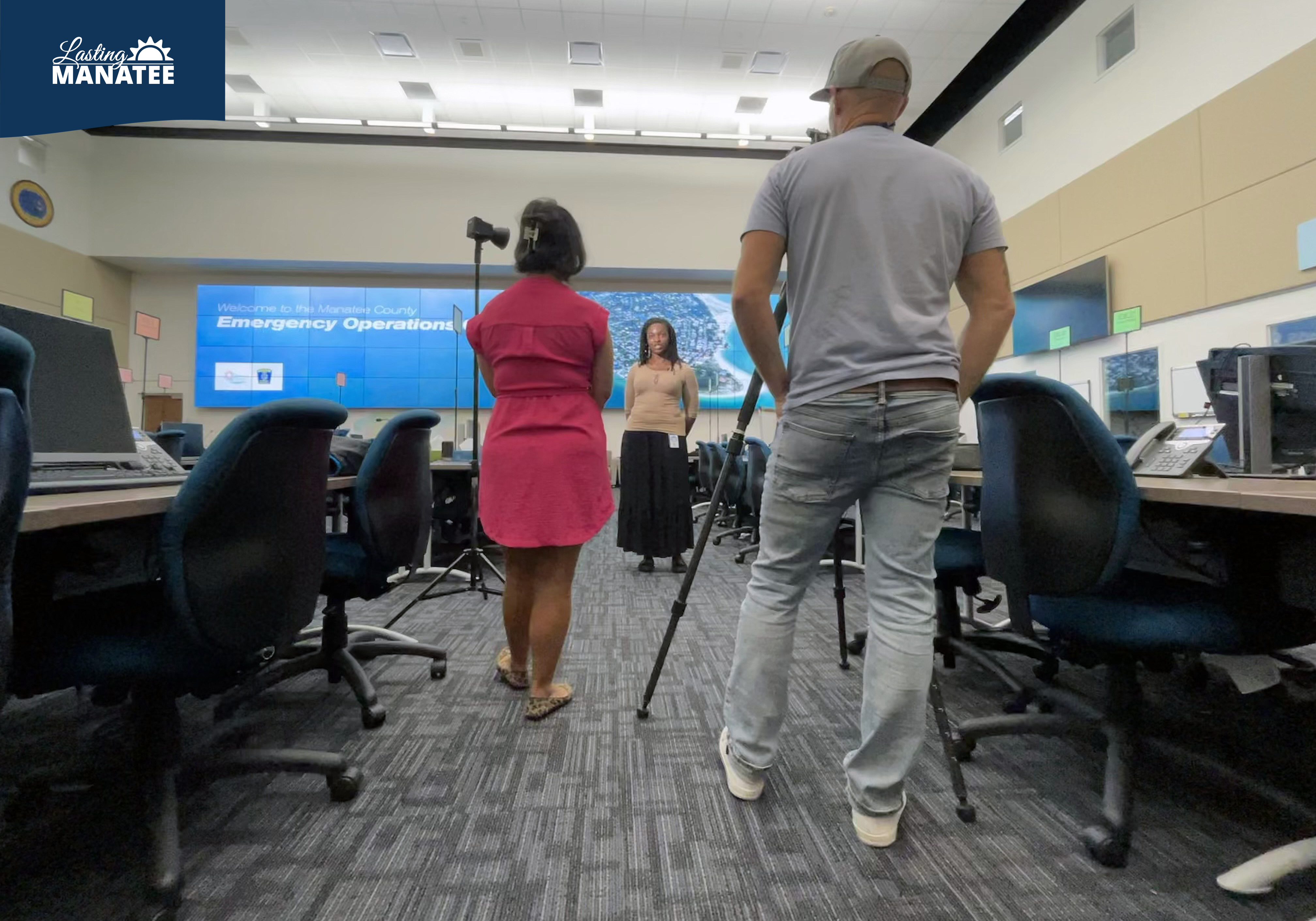 Lasting Manatee interviewed by FOX13 Kimberly Kuizon at the Emergency Operations Center Inside the Manatee County Emergency Operations Center, a staff member of Lasting Manatee speaks on camera while FOX13's reporter holds a microphone while their cameraman films. The room has desks and monitors, with a large TV screen on the back wall.